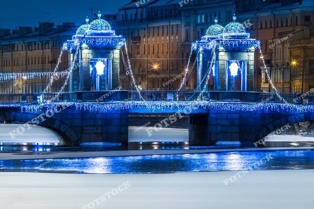 На фотографии изображён мост Ломоносова, раскинувшийся через реку Фонтанку в Санкт-Петербурге. Архитектурные башни моста украшены изысканной новогодней иллюминацией с голубыми гирляндами. Сияющие огни отражаются на замёрзшей поверхности реки, добавляя сцене сказочную атмосферу. Это один из самых известных мостов города, особенно впечатляющий в зимний праздничный сезон.
